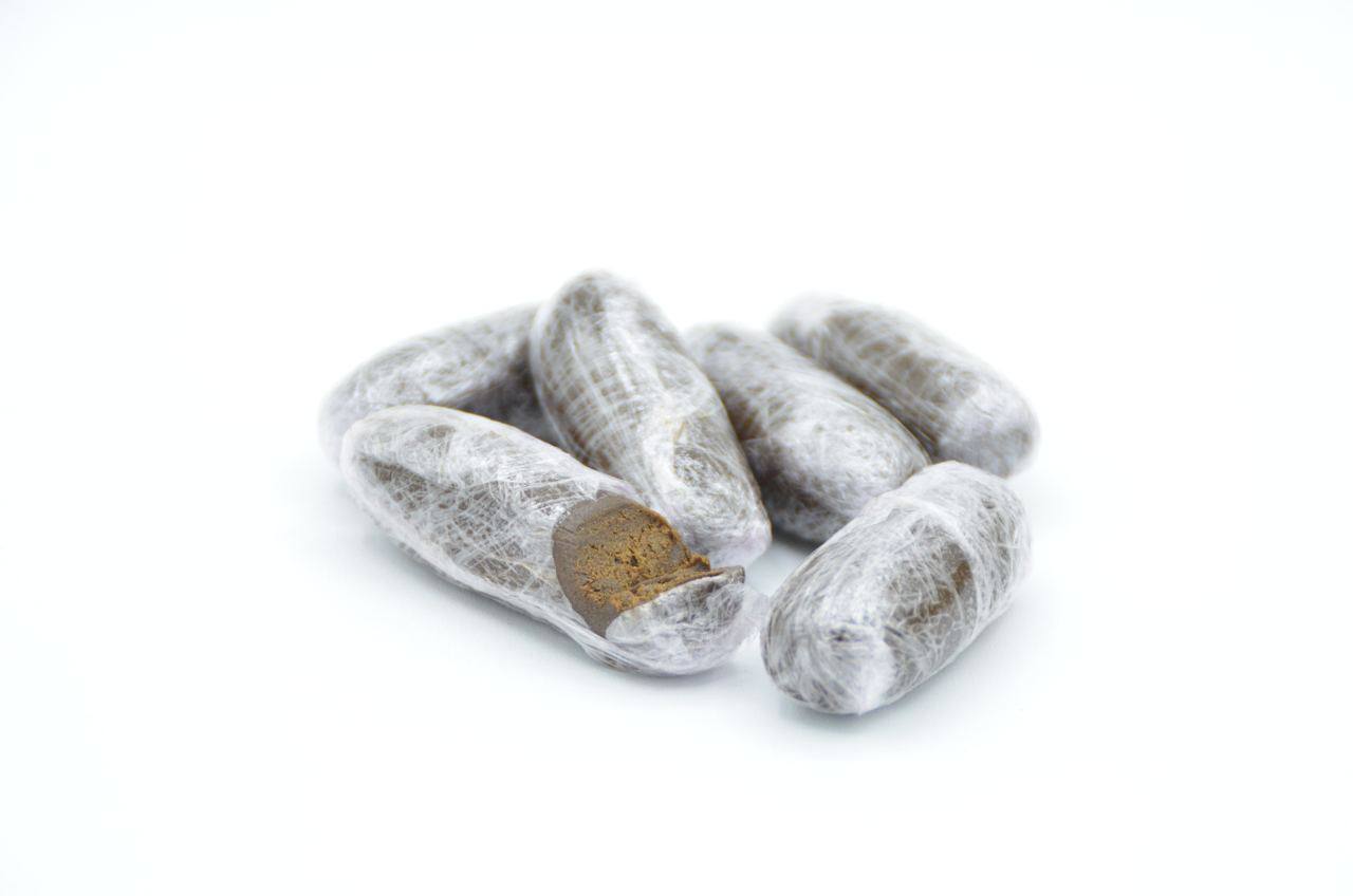 Plusieurs olivettes de hasch soigneusement emballées dans un film plastique transparent, dont l’une est partiellement ouverte, exposant son intérieur brun foncé et compact.