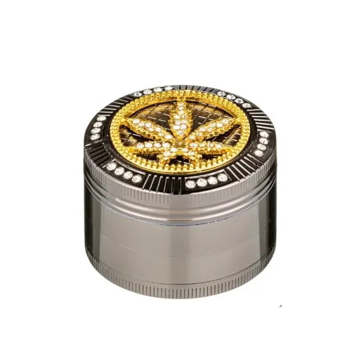 Grinder métallique argenté avec un couvercle orné d'une feuille de cannabis dorée en strass, entourée de pierres brillantes.